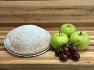 Round cake dusted with powdered sugar sits on a plate beside green apples and dark cherries on a wooden surface, a simple dessert still life