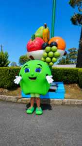 Green cartoon fruit mascot with a large fruit basket on a tall pole, standing outdoors on a sunny day.